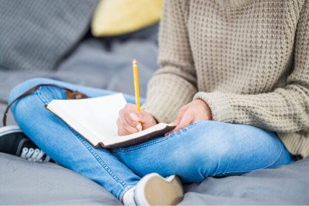 Vrouw zit op de bank in spijkerbroek met een schrift op schoot. Ze is aan het schrijven.