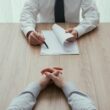 Twee mannen in pak zitten aan tafel in een serieus gesprek. De een houdt papieren vast en een pen.