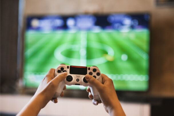 Twee handen houden een game controller vast. Op het beeldscherm zie je een voetbalspel.