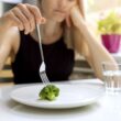 Jonge vrouw zit aan een eettafel met voor haar een bord. Er ligt 1 roosje broccoli op het bord.