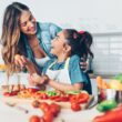 Een jonge vrouw en een meisje bereiden samen een gezonde maaltijd in de keuken. Ze kijken elkaar lachend aan.