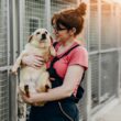 Vrouw houdt een puppy in haar armen bij een asiel. Op de achtergrond zie je de kooien van andere dieren.