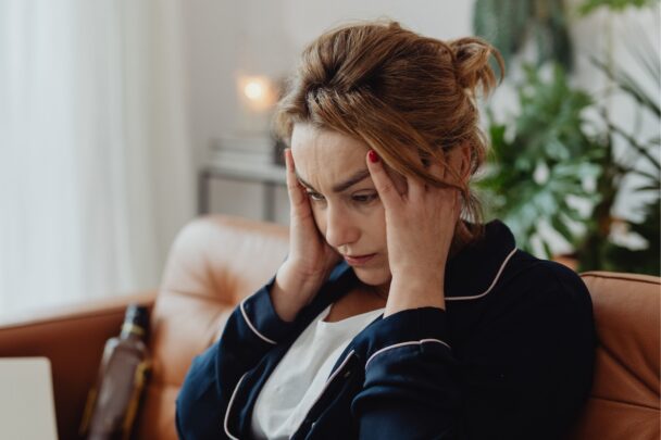 Vrouw zit met haar handen aan haar hoofd op de bank. Ze oogt gestresst.