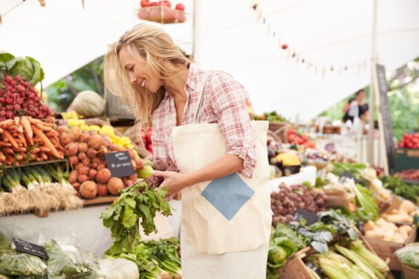 Blonde, jonge vrouw loopt met een canvas tas op een markt. Ze bekijkt groenten en fruit.