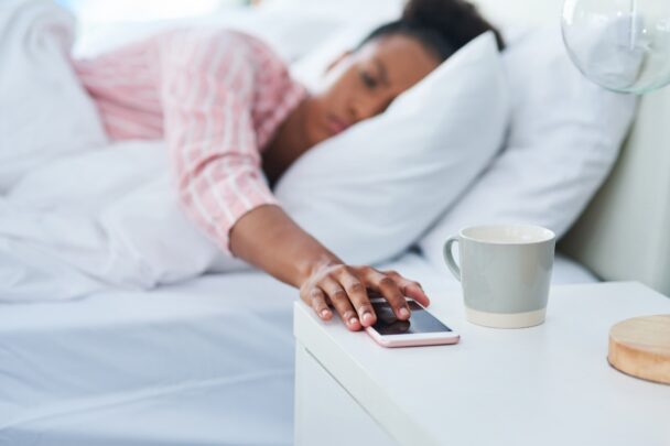 Vrouw met roze pyjama ligt in bed en drukt de wekker op haar nachtkastje uit. De wekker is een smarpthone.