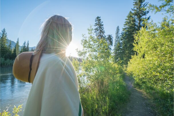 Achterkant van een vrouw met hoed die in een bosrijke omgeving loopt. De zon straalt en de lucht is blauw.