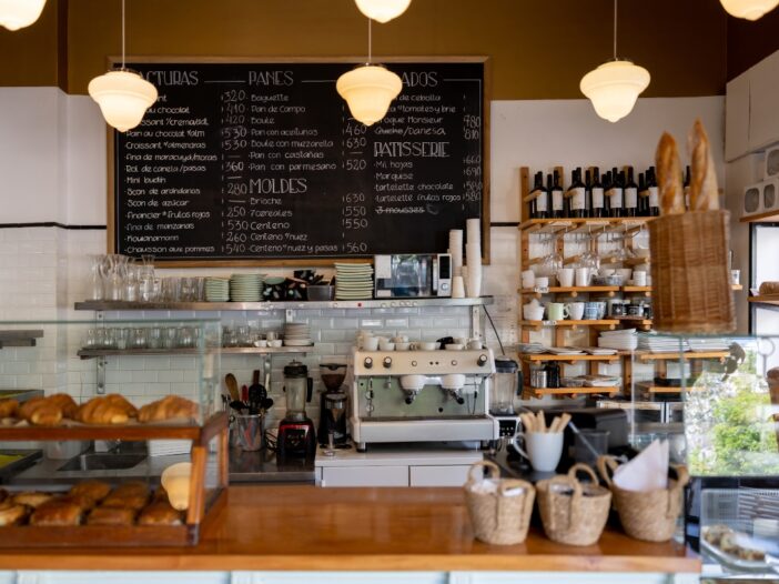 Toonbank van een café met allerlei kopjes erop, krijtborden met producten en prijzen aan de muur en een aantal hanglampen aan het plafond.