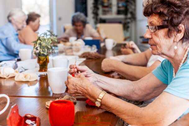 Vijf oudere dames zijn samen gezellig aan het haken en breien aan een grote tafel