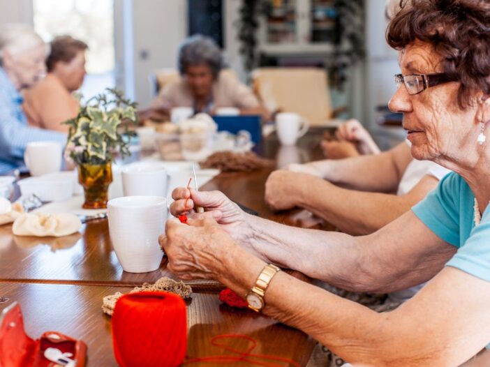 Vijf oudere dames zijn samen gezellig aan het haken en breien aan een grote tafel