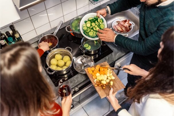 Drie mensen staan samen aan een fornuis een maaltijd met veel groenten te bereiden.