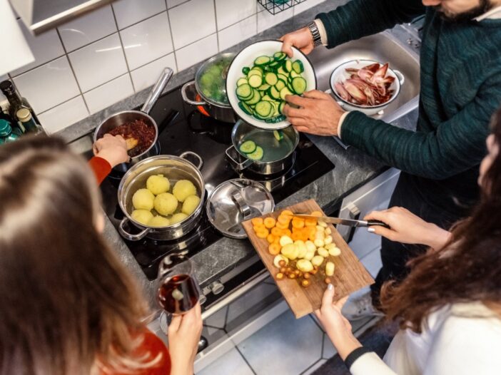 Drie mensen staan samen aan een fornuis een maaltijd met veel groenten te bereiden.