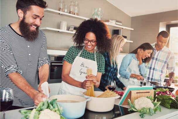 Drie vrouwen en twee mannen koken samen een maaltijd. De sfeer is gezellig.