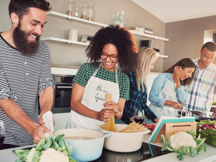 Drie vrouwen en twee mannen koken samen een maaltijd. De sfeer is gezellig.