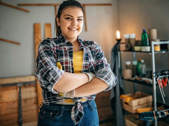 Jonge vrouw kijkt lachend in de camera met haar armen over elkaar. Ze oogt trots. Op de achtergrond zien we wat houten planken en gereedschap in een stellingkast.