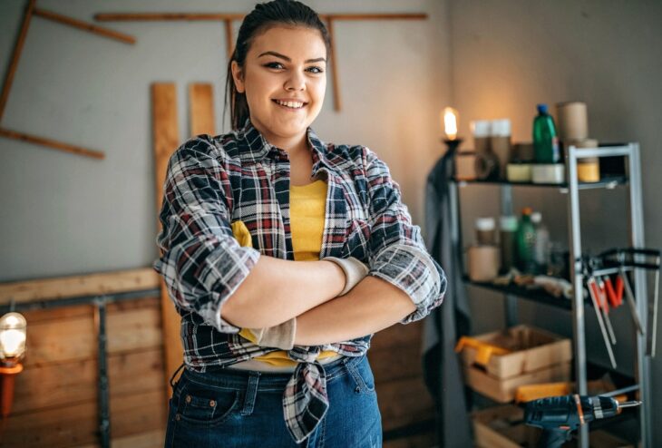 Jonge vrouw kijkt lachend in de camera met haar armen over elkaar. Ze oogt trots. Op de achtergrond zien we wat houten planken en gereedschap in een stellingkast.