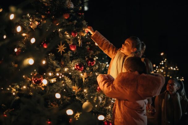 Twee meisjes versieren een kerstboom buiten in het donker. Hun moeder zit er gehurkt bij.