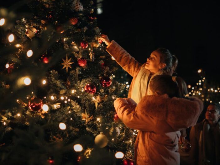 Twee meisjes versieren een kerstboom buiten in het donker. Hun moeder zit er gehurkt bij.