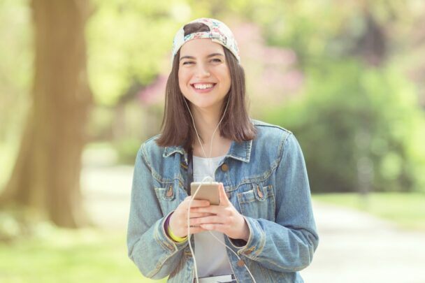 Jonge meid met pet en smartphone in haar handen kijkt blij in de camera.