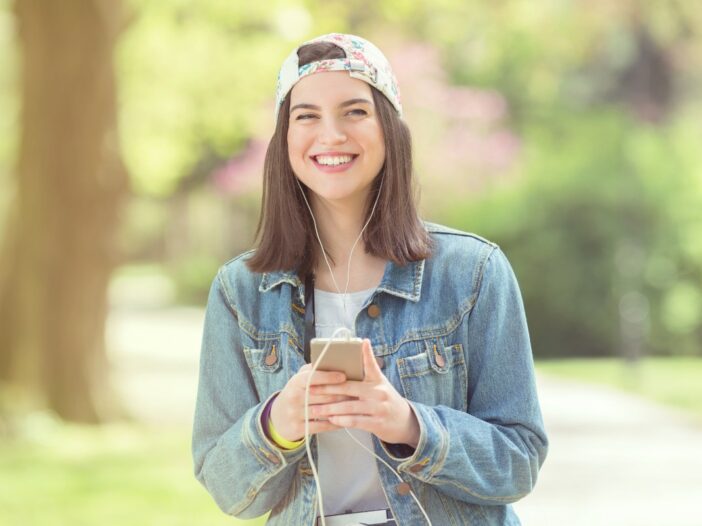 Jonge meid met pet en smartphone in haar handen kijkt blij in de camera.