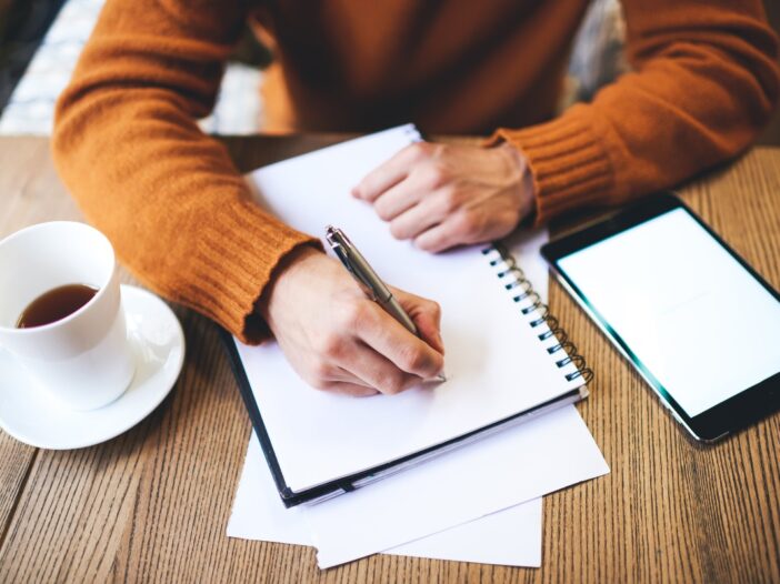 Man schrijft in een notitieboek. Er staat een kopje koffie op tafel.