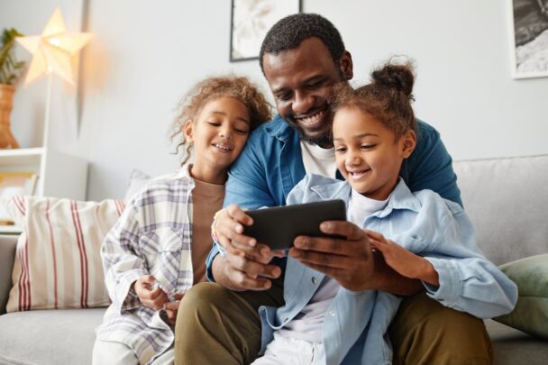 Vader zit met zijn twee dochters op de bank en ze kijken allemaal naar een smartphone. Ze ogen vrolijk.