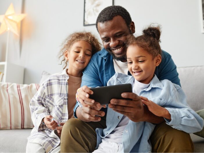 Vader zit met zijn twee dochters op de bank en ze kijken allemaal naar een smartphone. Ze ogen vrolijk.