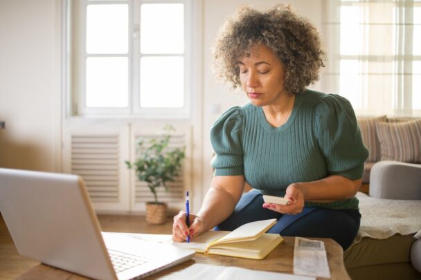 Vrouw zit op de bank en doet administratieve klusjes op haar smartphone, laptop en op papier.