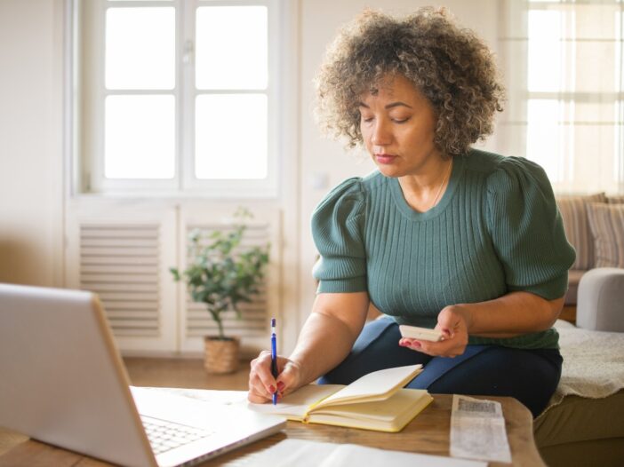 Vrouw zit op de bank en doet administratieve klusjes op haar smartphone, laptop en op papier.