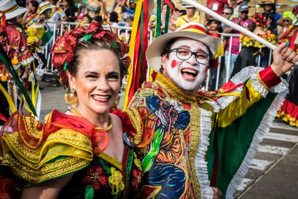 Man en vrouw in kleurrijke carnavalskleding kijken blij de camera in.