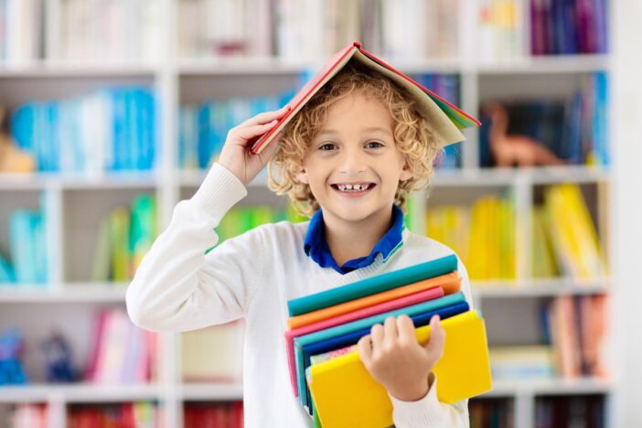 Jongen van basisschoolleeftijd heeft schoolboeken in zijn handen en een boek op zijn hoofd. Hij lacht breed.