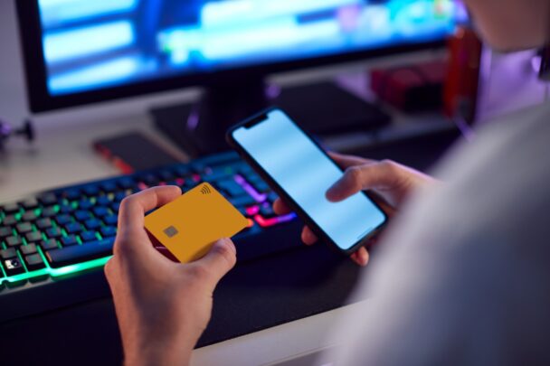 Man achter computerscherm logt op zijn smartphone in en houdt een creditcard in zijn handen.