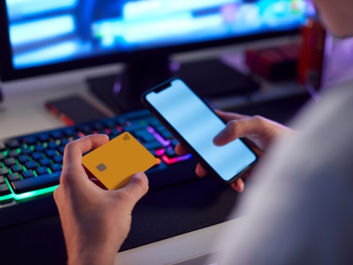 Man achter computerscherm logt op zijn smartphone in en houdt een creditcard in zijn handen.