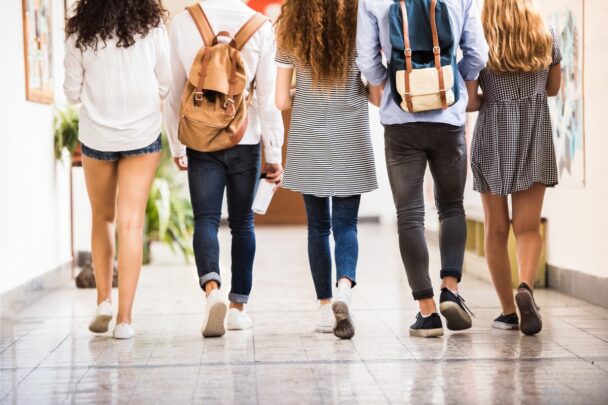 Vijf tieners lopen door een gang in een schoolgebouw. Een paar dragen er een rugzak op hun rug.