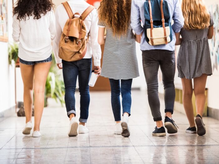 Vijf tieners lopen door een gang in een schoolgebouw. Een paar dragen er een rugzak op hun rug.