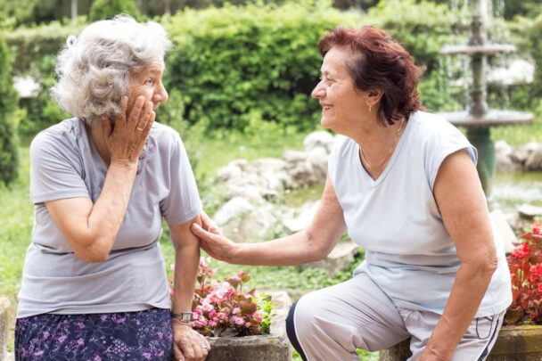 Twee oudere dames zijn samen in gesprek. Ze zitten buiten op een bankje.