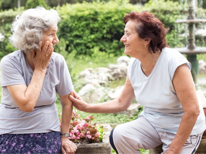 Twee oudere dames zijn samen in gesprek. Ze zitten buiten op een bankje.