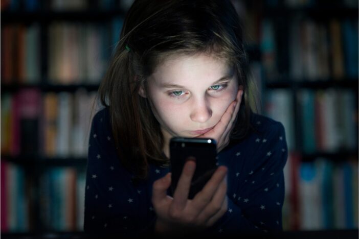 Meisje zit in het donker en staart naar het oplichtende scherm van haar smartphone.