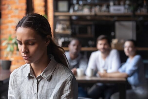 Jonge vrouw kijkt terneergeslagen. Op de achtergrond zie je een groepje mensen zitten wat naar haar staart.