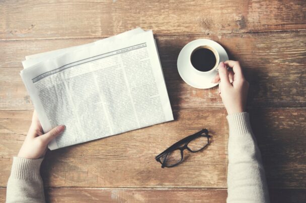 Tafel met daarop een bril, kop koffie en handen die een krant vasthouden.