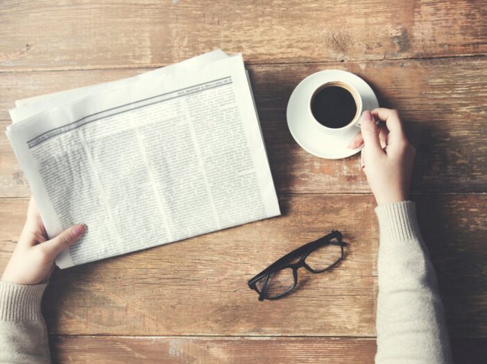 Tafel met daarop een bril, kop koffie en handen die een krant vasthouden.