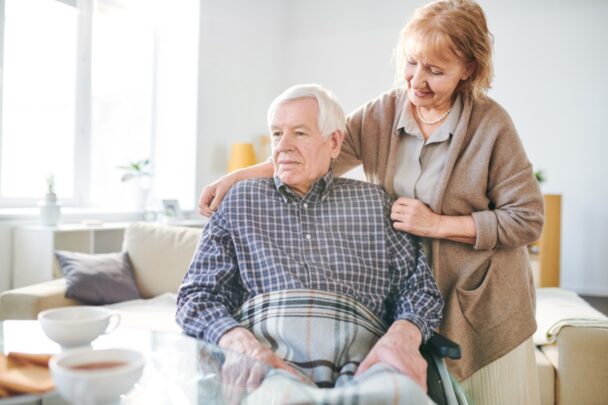 Oudere vrouw houdt zorgzaam de schouders van een oudere man vast.